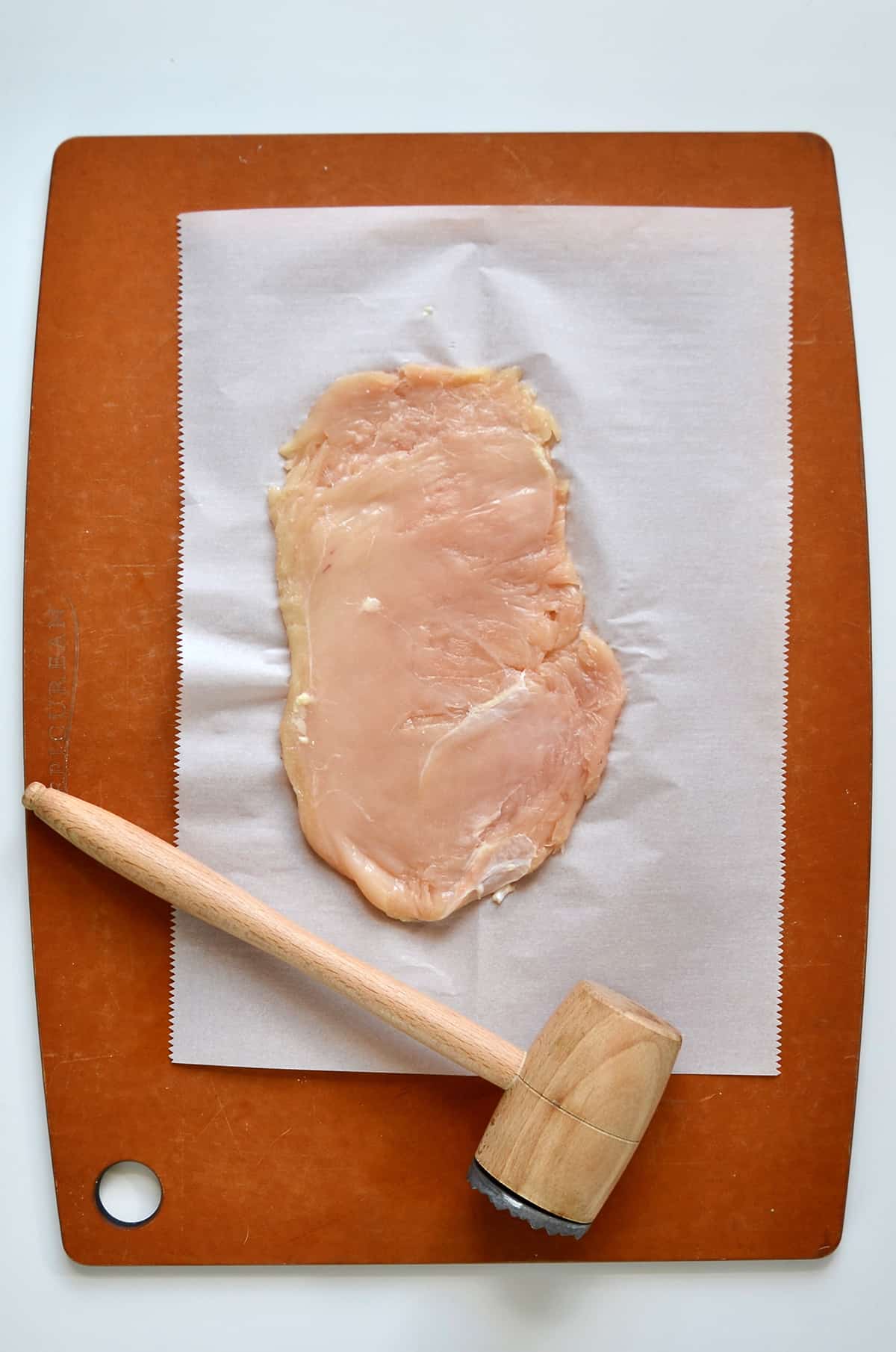 A piece of thinly pounded chicken is on a piece of parchment paper on a cutting board, with a wooden meat mallet nearby.