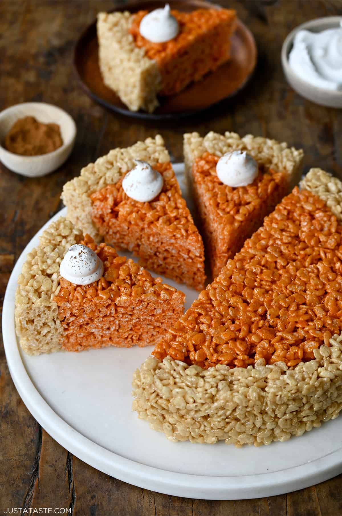 Pumpkin-flavored Rice Krispies treats cut into pie wedges and topped with dollops of whipped cream on a round serving platter next to a small bowl containing pumpkin pie spice.