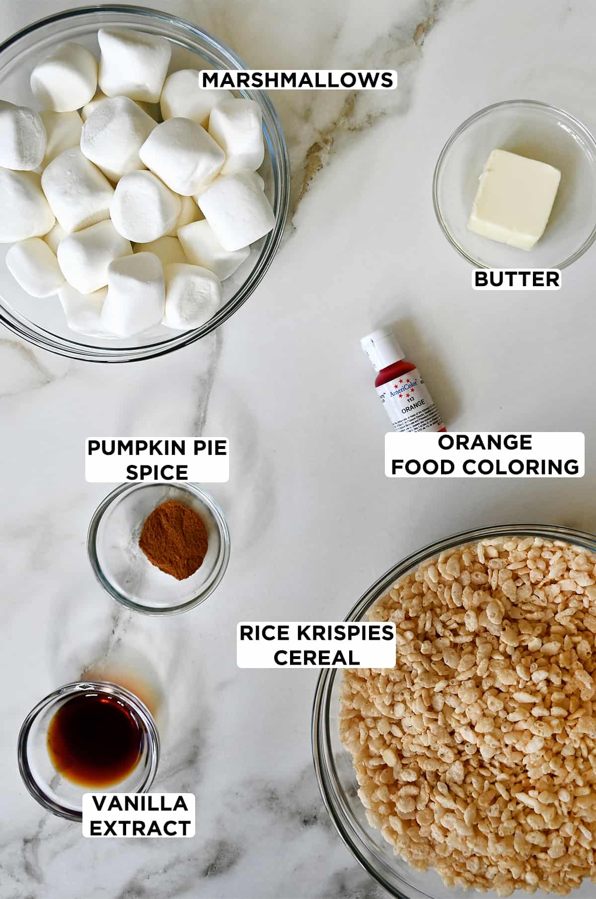 Clear bowls containing marshmallows, butter, Rice Krispies cereal, pumpkin pie spice and vanilla extract next to a bottle of orange food coloring.