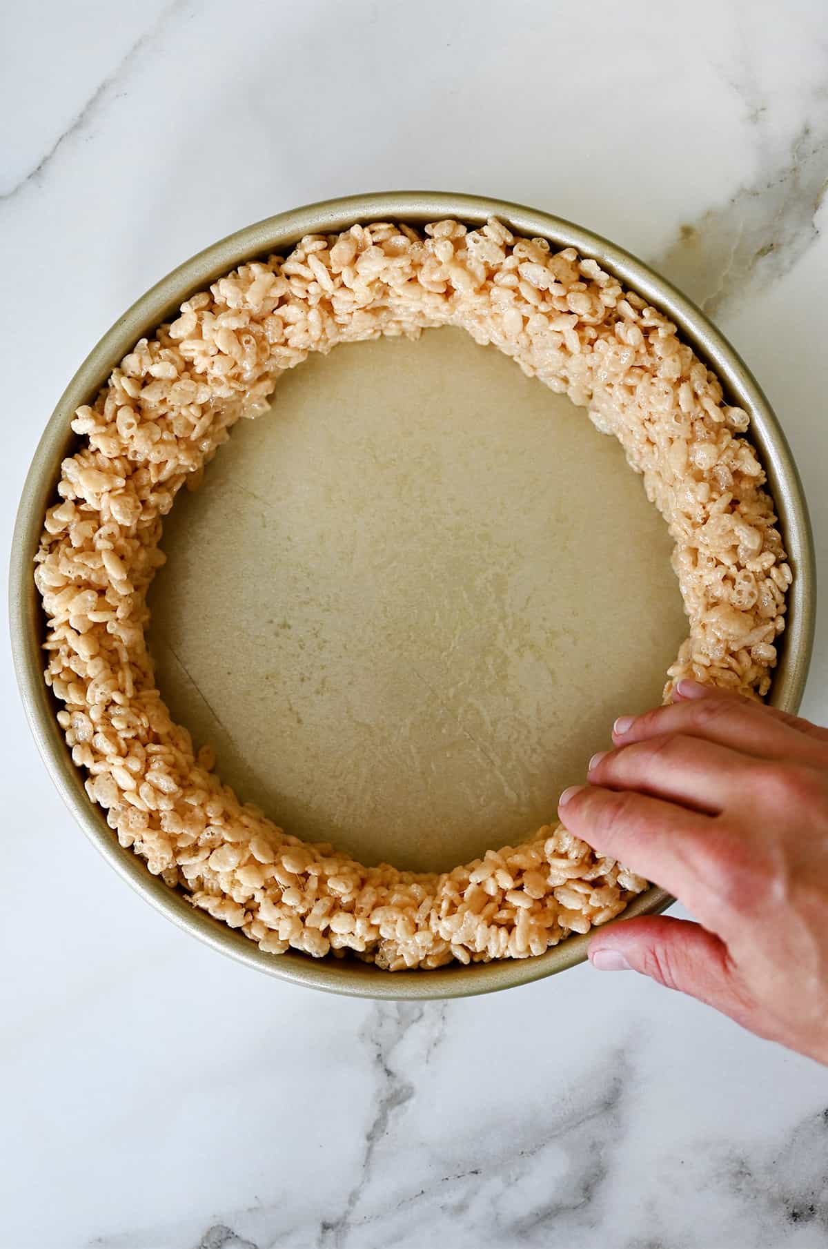 A hand molds vanilla marshmallow treats around the edge of a cake pan to form a "crust." 