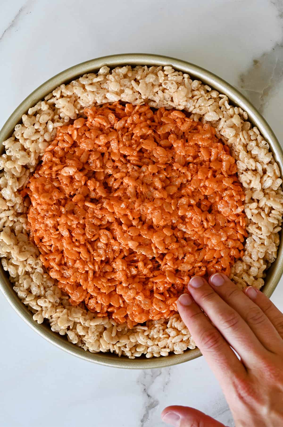 A hand gently presses the pumpkin marshmallow treats into the center of a round cake pan.