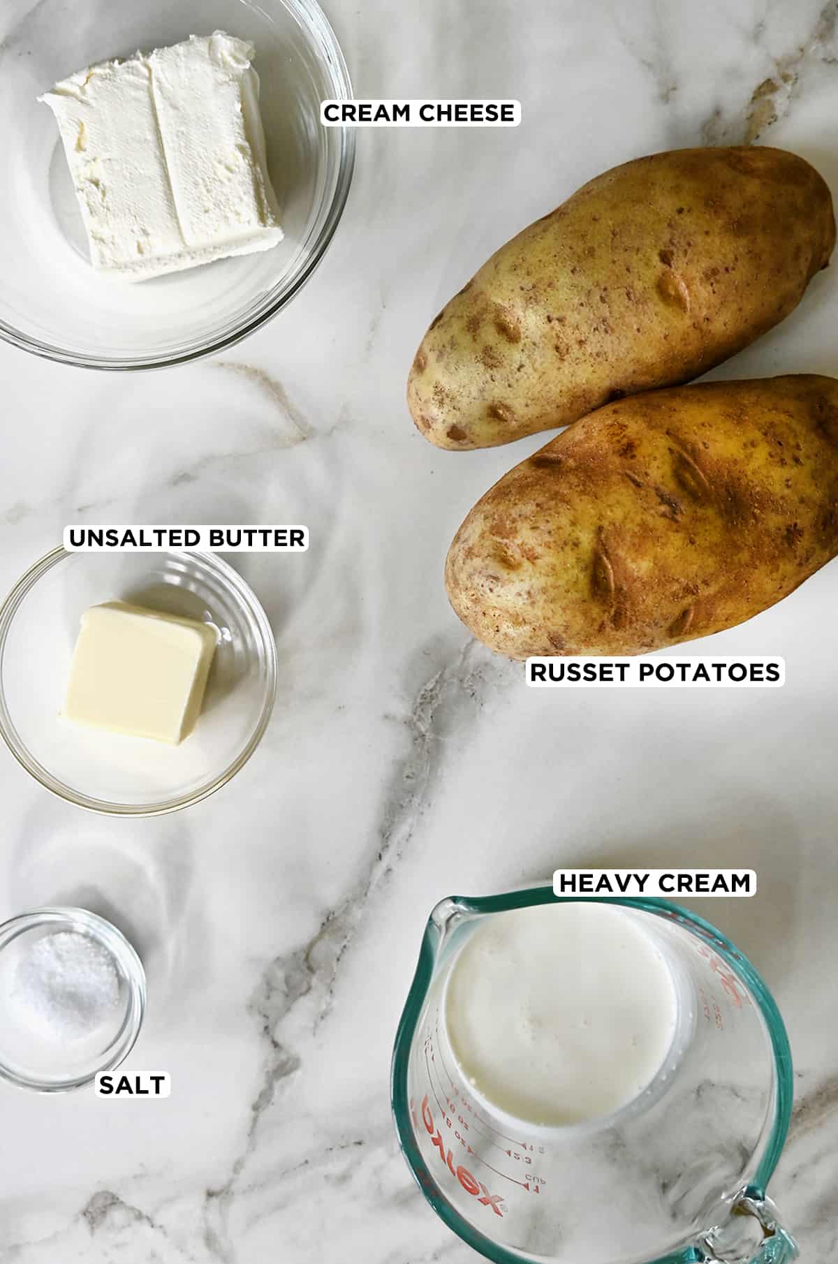 Two Russet potatoes next to various sizes of clear bowls containing cream cheese, butter, heavy cream and salt.
