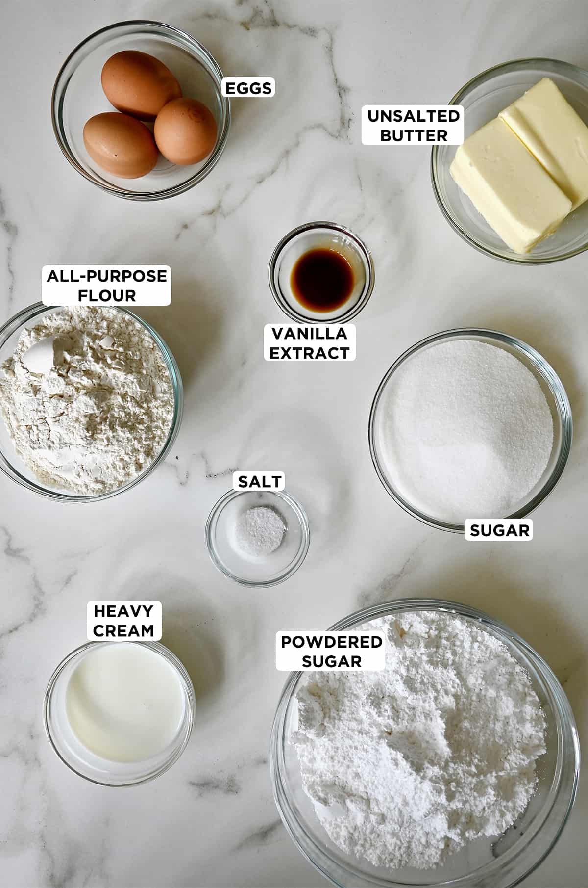 Various sizes of clear bowls containing two eggs, vanilla extract, butter, sugar, salt, powdered sugar, heavy cream and all-purpose flour.