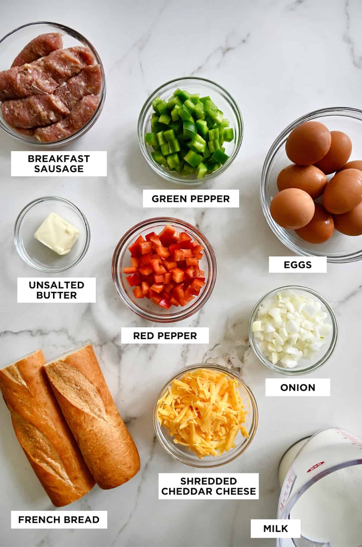 A labeled image of all the ingredients needed to make an egg casserole, including breakfast sausage, green and red bell peppers, eggs, butter, onion, cheddar cheese, milk and a loaf of French bread.