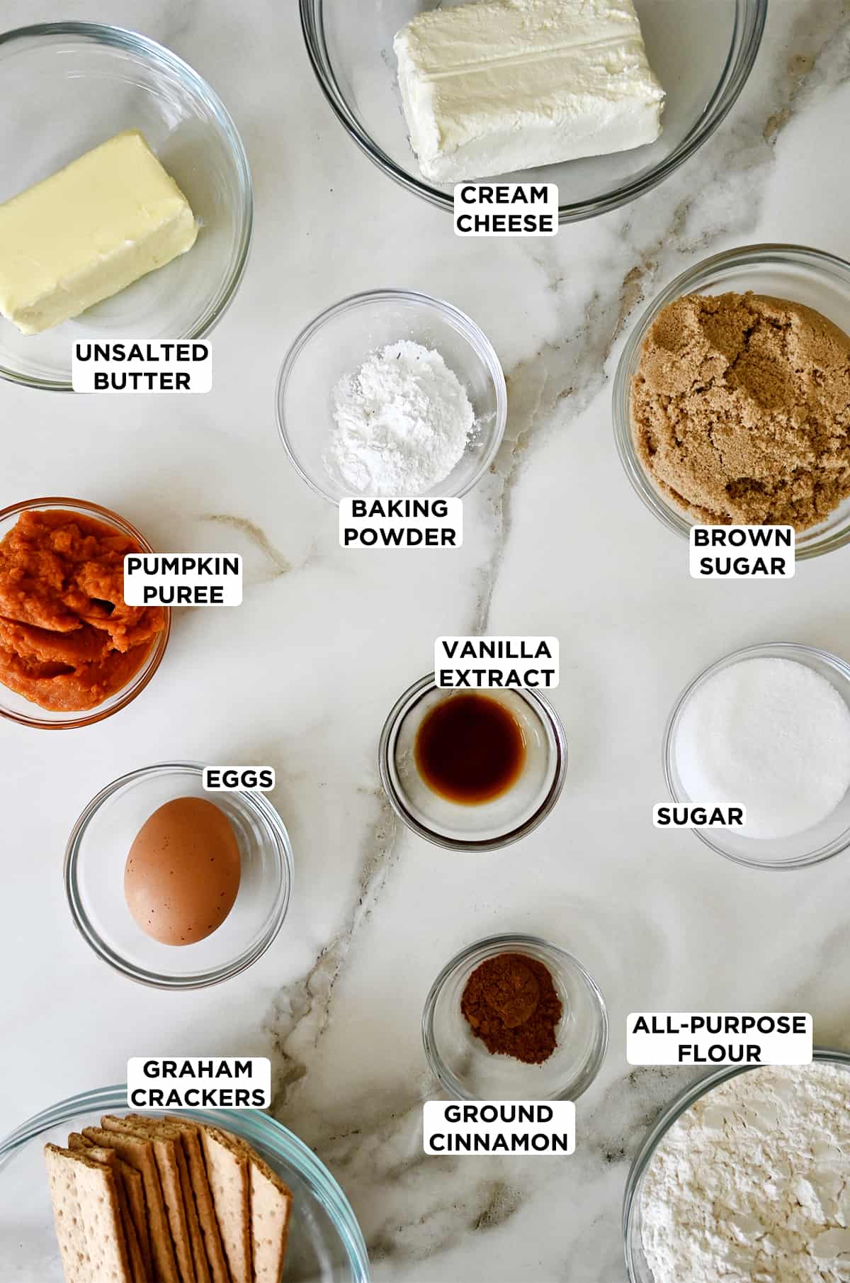 Various sizes of clear bowls containing all of the ingredients needed to make pumpkin cheesecake cookies: cream cheese, brown sugar, white sugar, flour, ground cinnamon, vanilla extract, baking powder, butter, pumpkin puree, an egg and graham crackers.