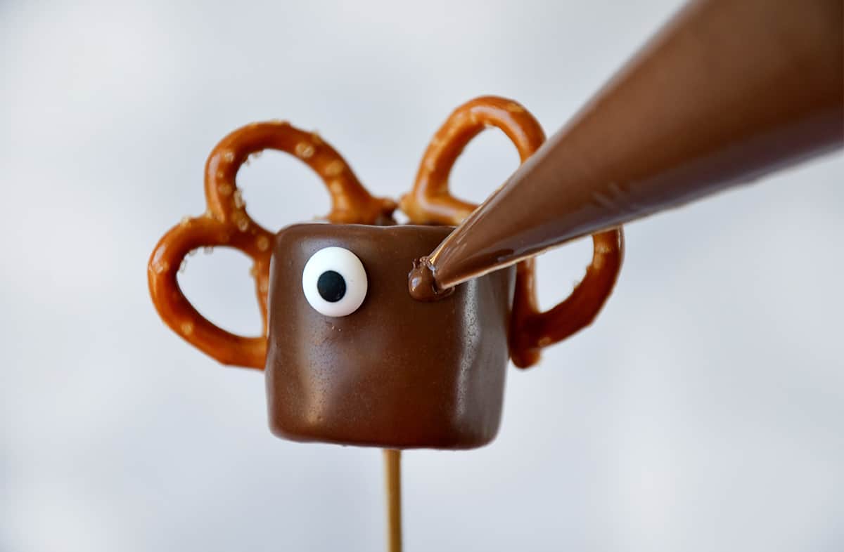 Melted chocolate from a piping bag is being applied to a reindeer pop to be the "glue" for a candy eye.