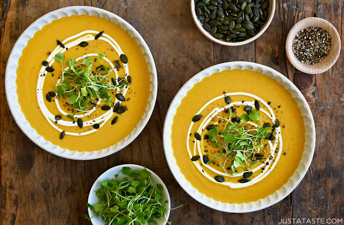 Two bowls containing coconut pumpkin soup garnished with micro greens, toasted pumpkin seeds and a drizzle of sour cream.