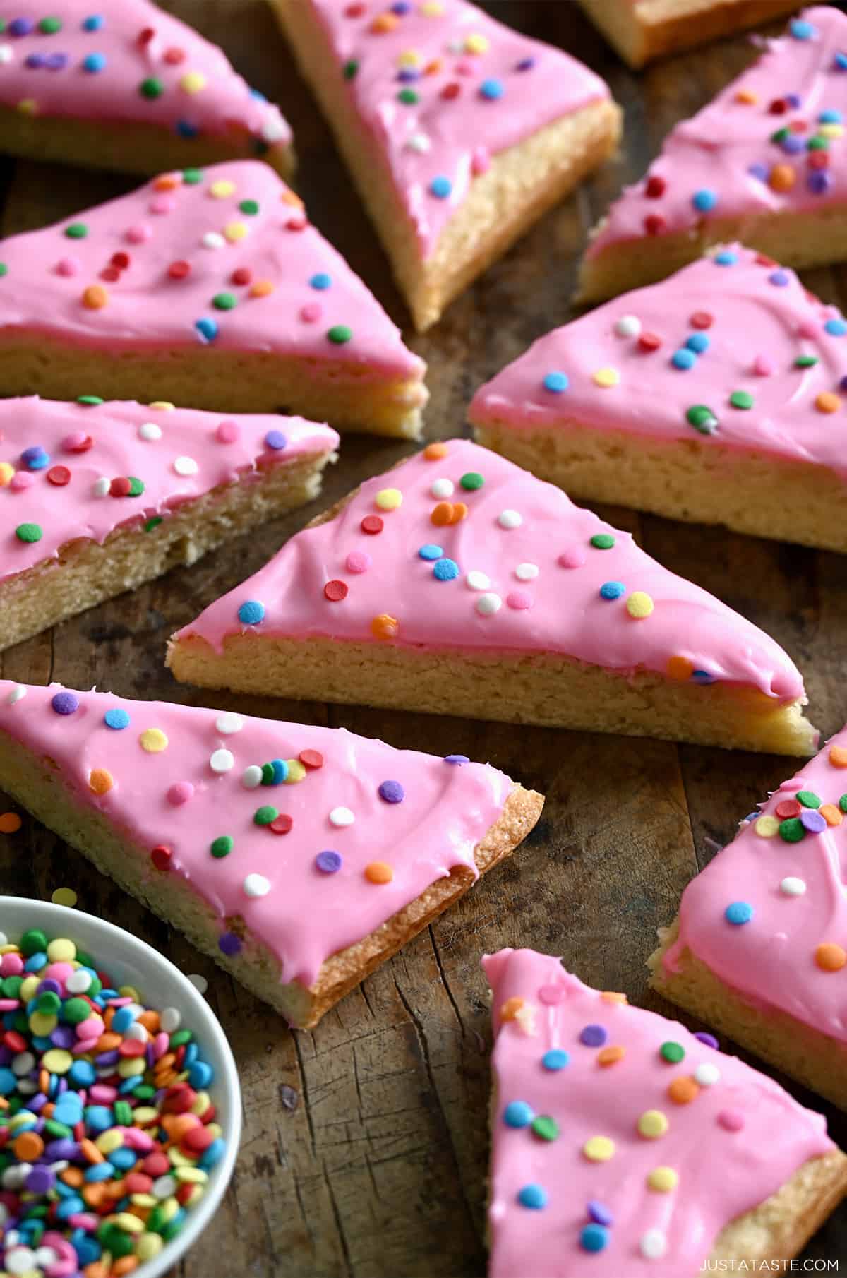 Sugar cookie bars with pink frosting and sprinkles on a wooden surface next to a small bowl containing rainbow sprinkles.