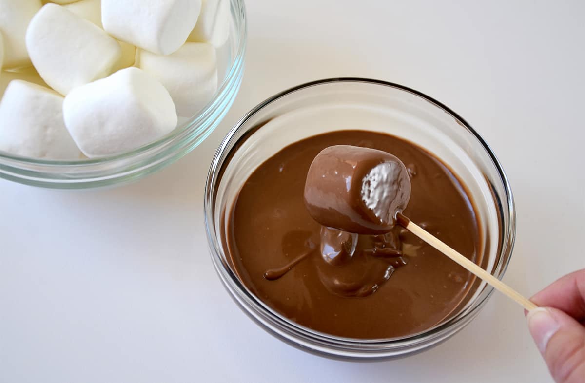 A marshmallow on a wooden skewer being dipped into a bowl containing melted chocolate. Another bowl containing marshmallows is next to it.