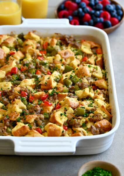 An egg casserole with breakfast sausage and colorful bell peppers in a white baking dish next to a bowl of fresh berries.