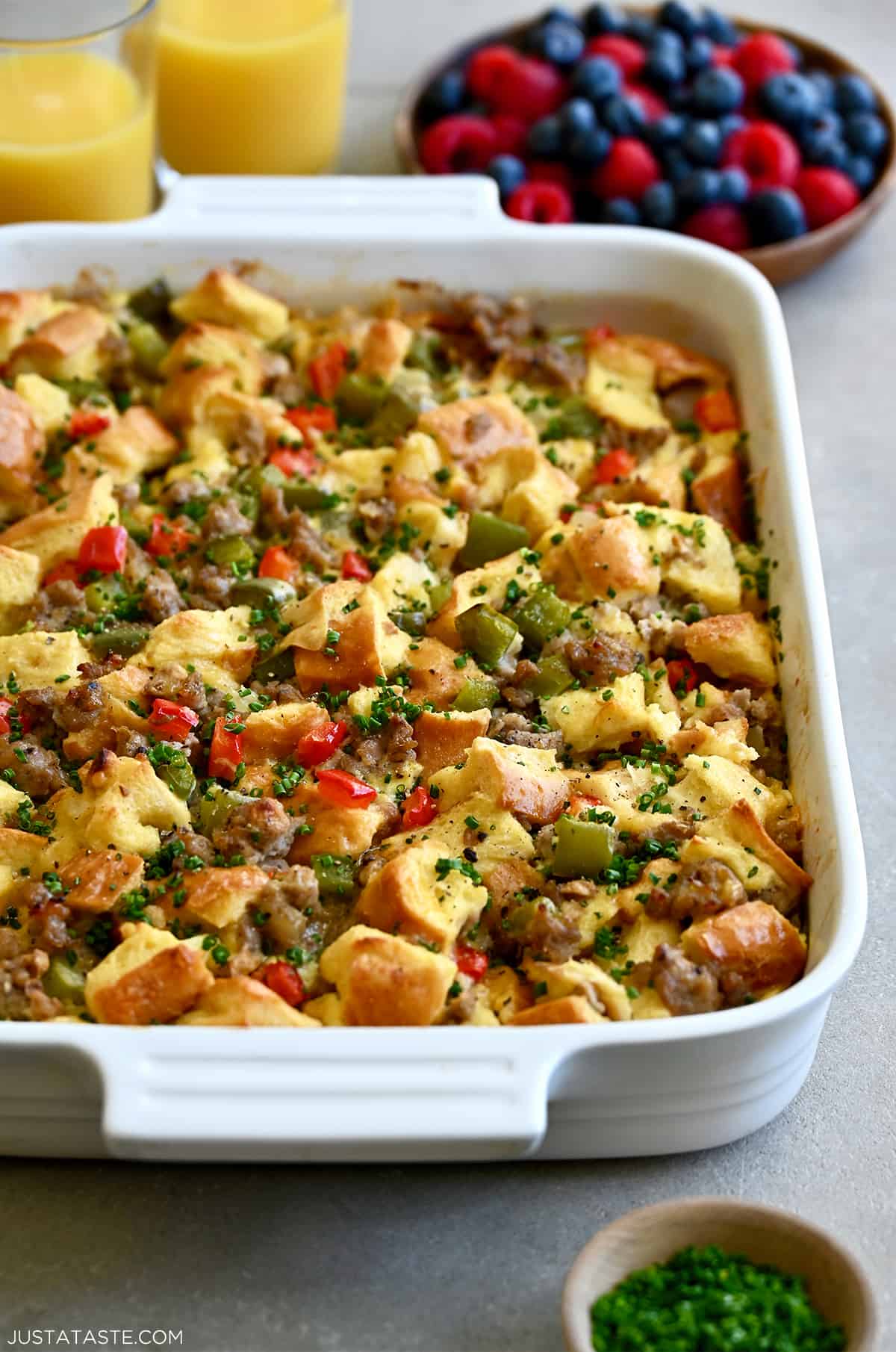 An egg casserole with breakfast sausage and colorful bell peppers in a white baking dish next to a bowl of fresh berries.