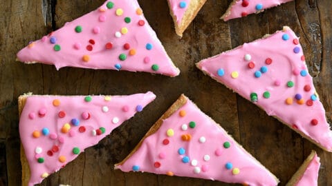 Pink-frosted sugar cookie bars with rainbow sprinkles next to a small bowl containing sprinkles.