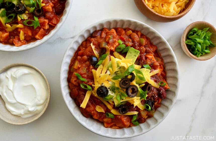 A bowl containing 30-Minute Leftover Turkey Chili topped with sour cream, diced avocado, black olives and shredded cheddar cheese next to a small bowl with sour cream