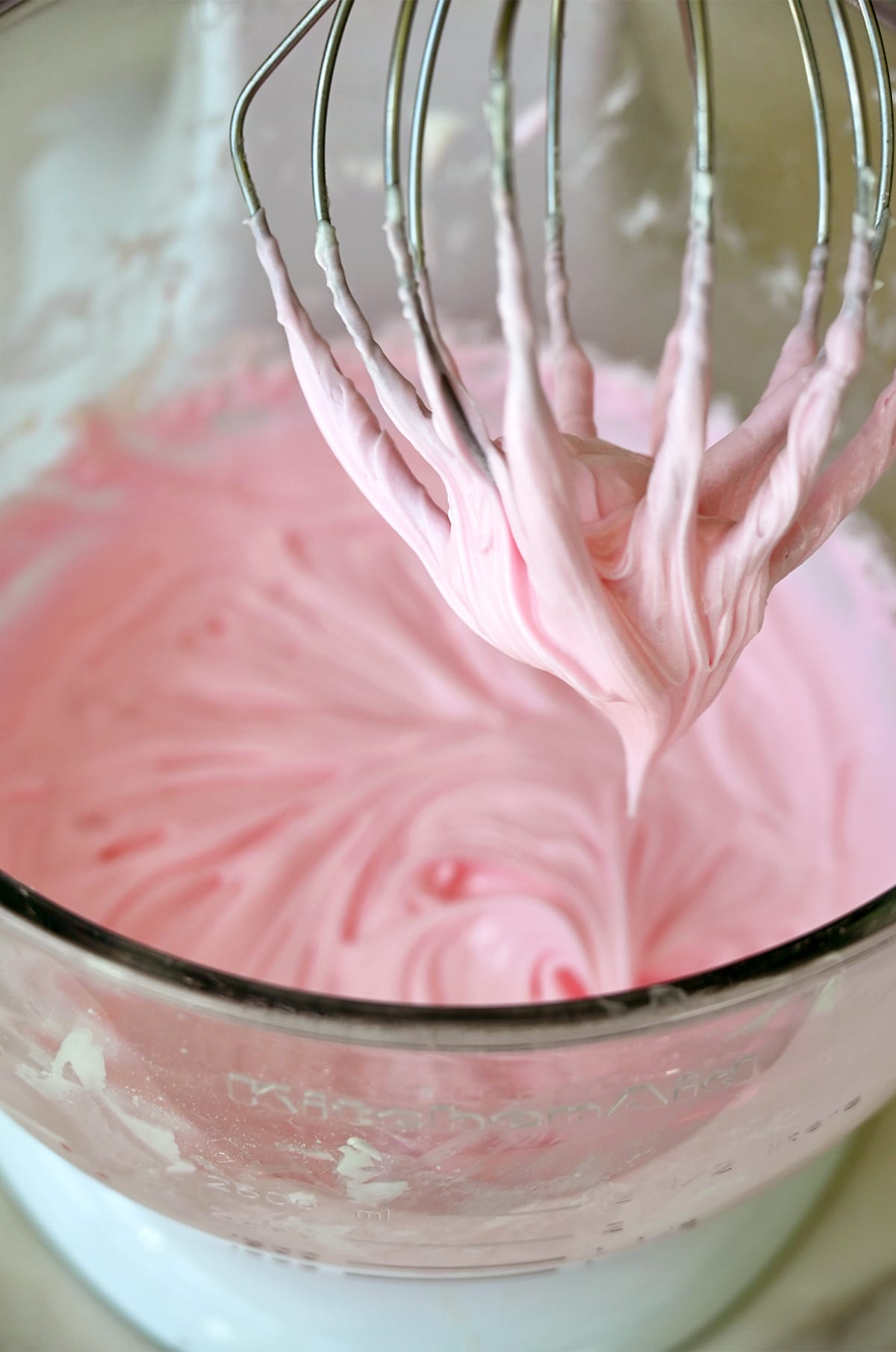 Pink buttercream frosting in a stand mixer bowl with the whisk attachment.