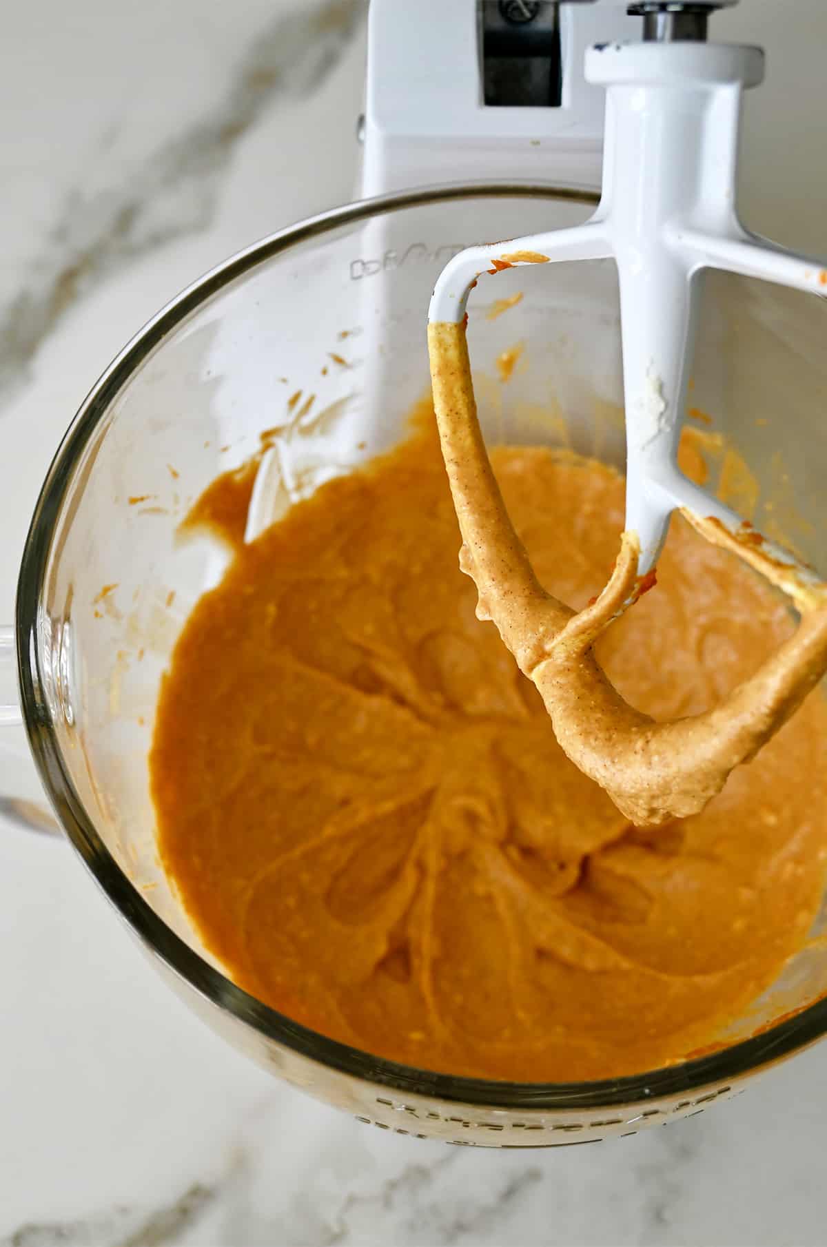 A paddle attachment of a stand mixer hovers over a stand mixer bowl containing pumpkin cheesecake mixture.