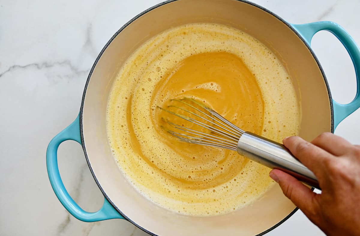 Coconut milk being whisked into a pumpkin soup base in a large stockpot.