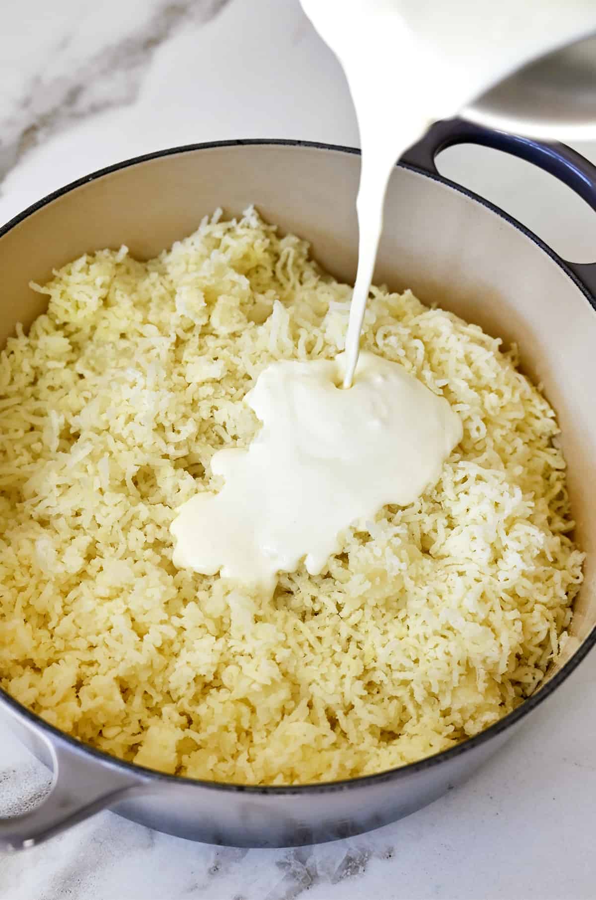 Riced potatoes in a large stockpot with heavy cream being poured into the pot.