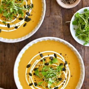 Two bowls containing Thai coconut pumpkin soup garnished with pumpkin seeds, micro greens and a drizzle of sour cream.
