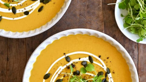 Two bowls containing Thai coconut pumpkin soup garnished with pumpkin seeds, micro greens and a drizzle of sour cream.