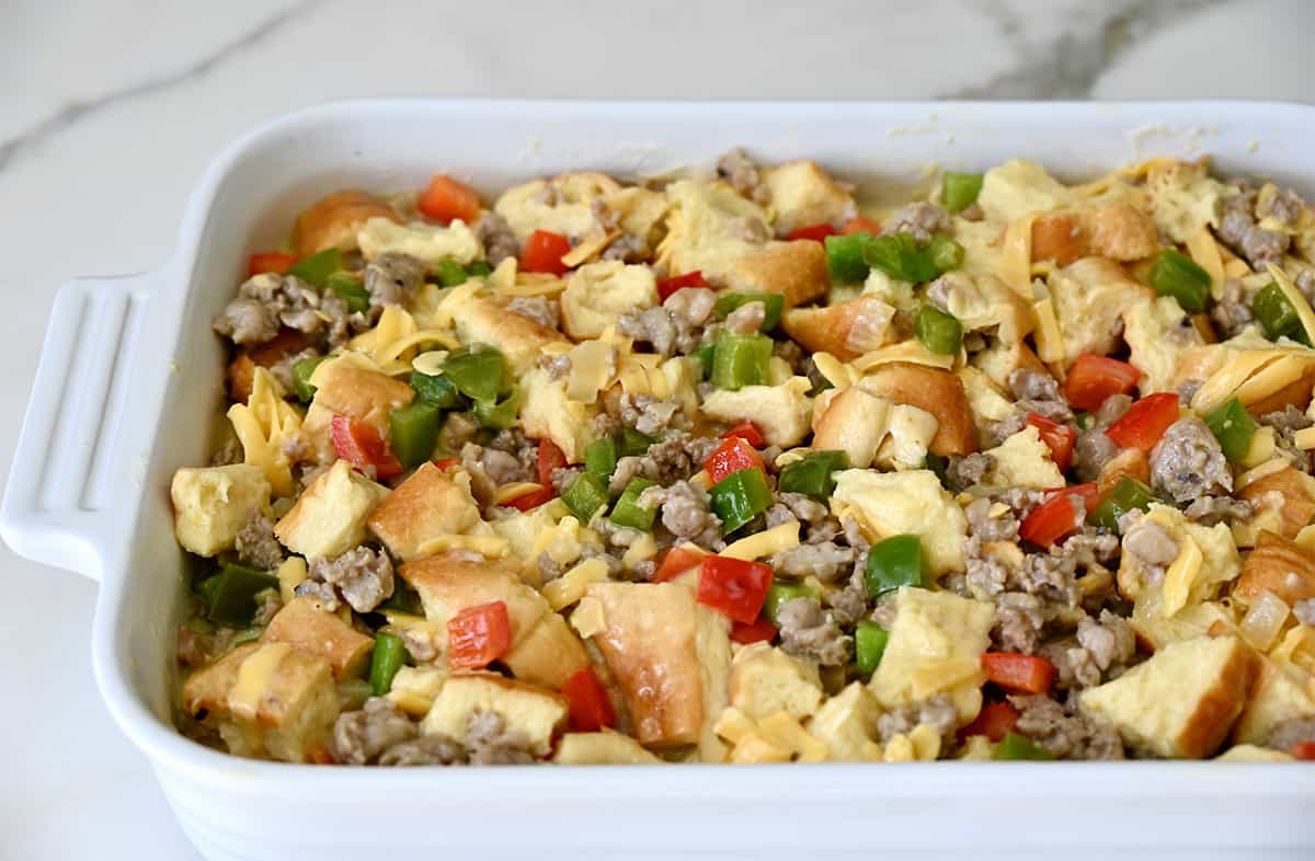 Cubed bread in an egg mixture with breakfast sausage and diced peppers in a white baking dish.