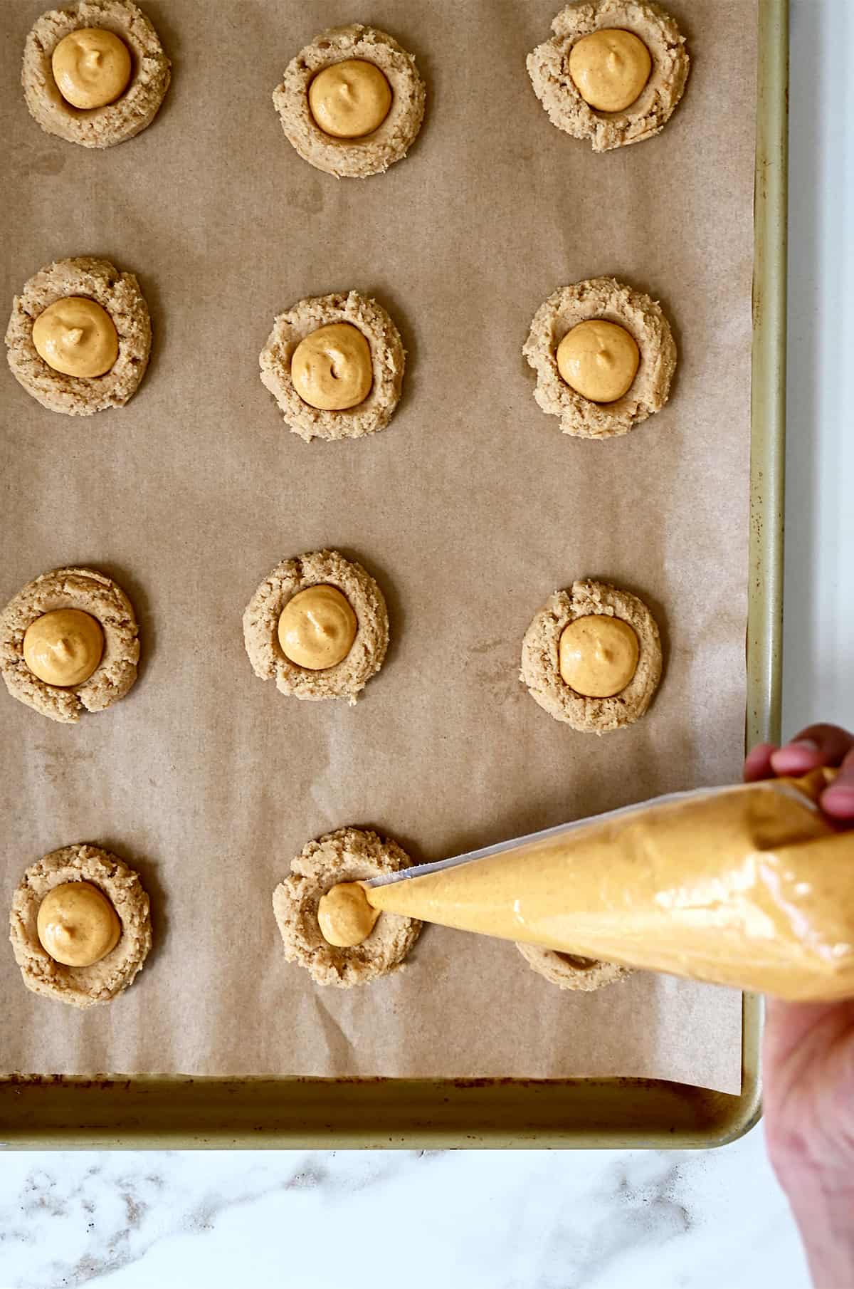 A hand holding a plastic piping bag pipes pumpkin cheesecake into the center of cookie dough on a parchment paper-lined baking sheet.