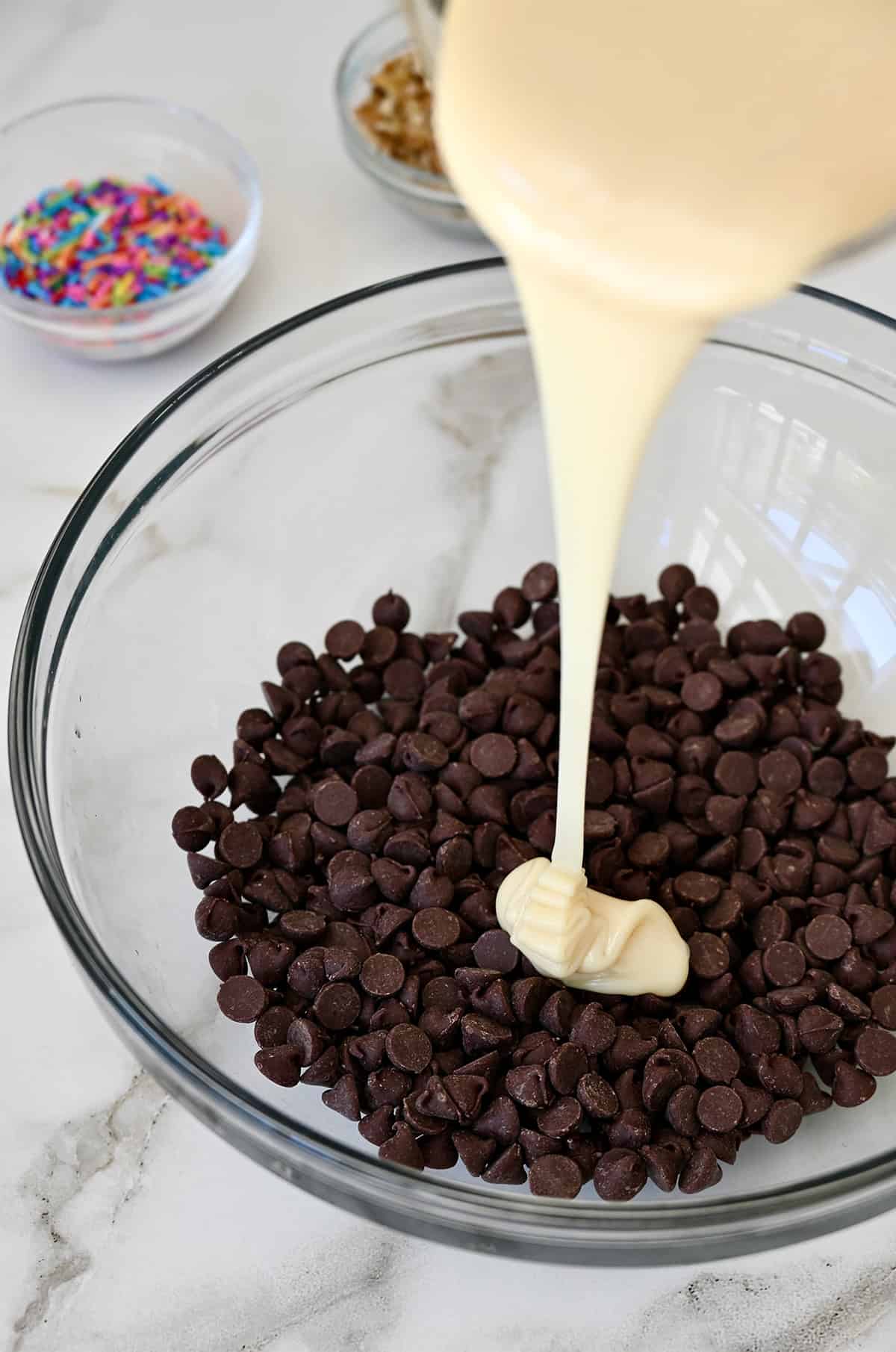 Sweetened condensed milk being poured over chocolate chips in a glass bowl.