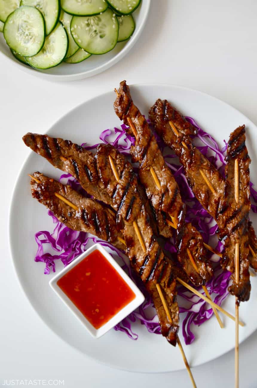 A white plate containing 5-Ingredient Beef Satays over a bed of shredded cabbage next to a small ramekin containing sweet chili sauce.