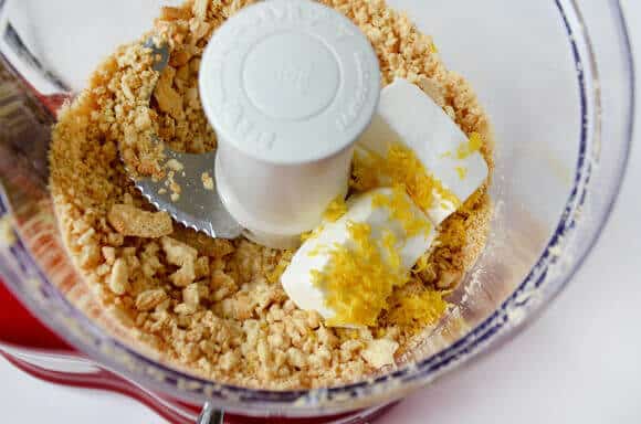 A top-down view of blended vanilla cookies, cream cheese and lemon zest in the bowl of a food processor