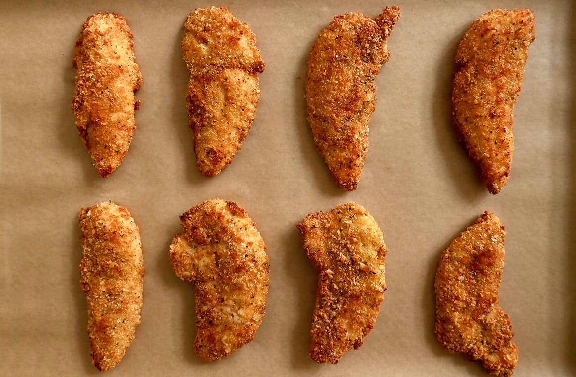 Two rows of Parmesan crusted chicken tenders on a parchment paper-lined baking sheet