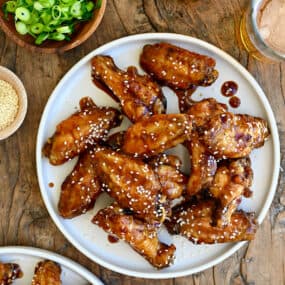 Teriyaki chicken wings garnished with sesame seeds on a white plate next to two glasses containing beer and a small bowl with sliced scallions.