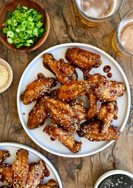 Teriyaki chicken wings garnished with sesame seeds on a white plate next to two glasses containing beer and a small bowl with sliced scallions.