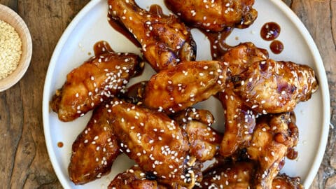 Teriyaki chicken wings garnished with sesame seeds on a white plate next to two glasses containing beer and a small bowl with sliced scallions.