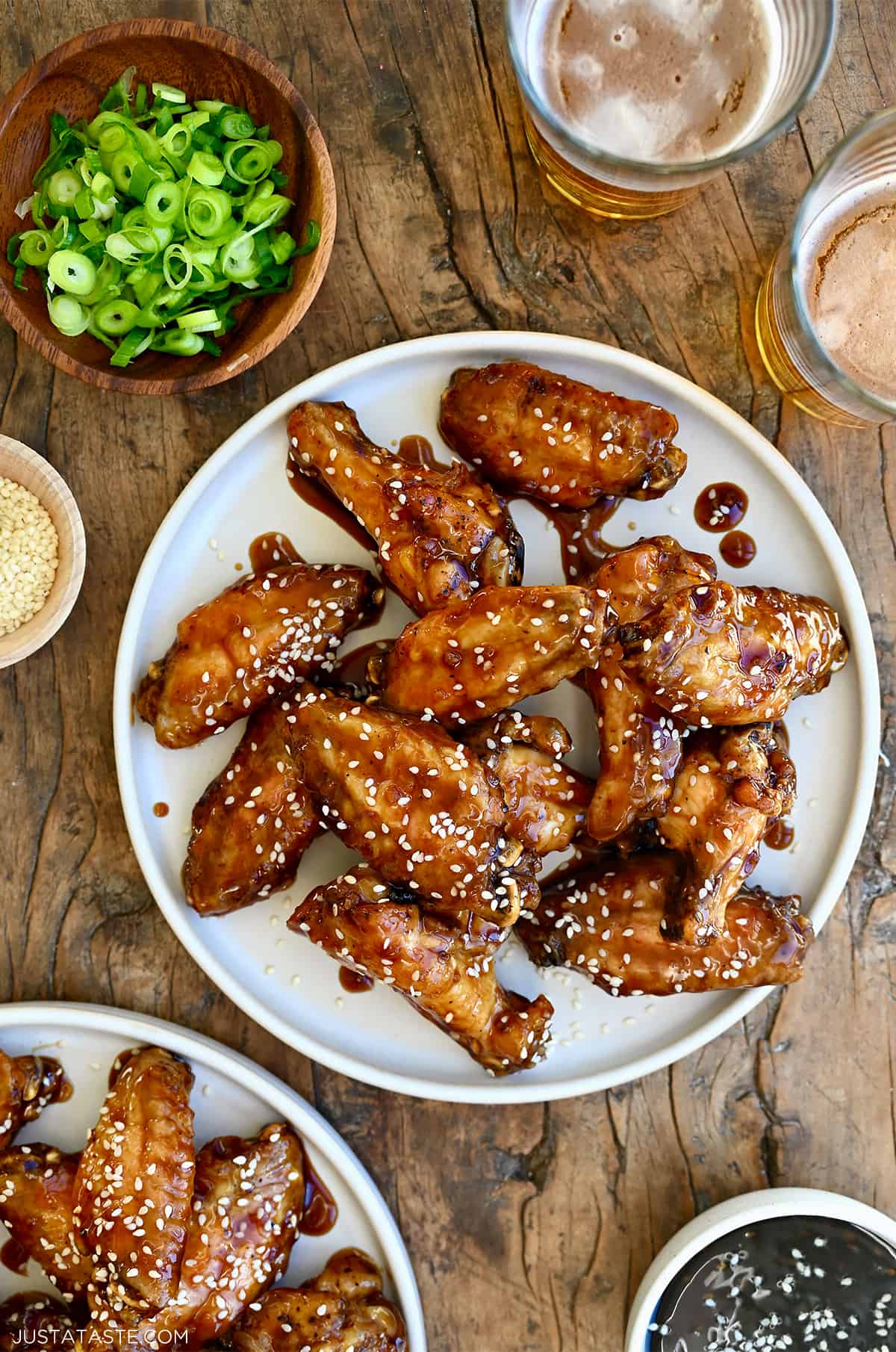 Teriyaki chicken wings garnished with sesame seeds on a white plate next to two glasses containing beer and a small bowl with sliced scallions.