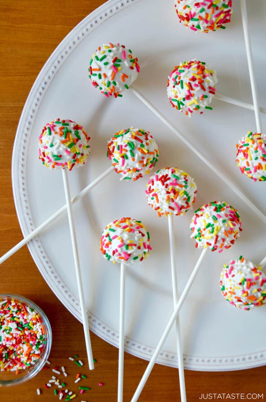 A top-down view of No-Bake Cheesecake Cookie Pops studded with rainbow sprinkles on a white plate next to a small bowl containing rainbow sprinkles