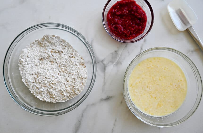 A top-down view of three bowls containing wet and dry ingredients and mashed berries