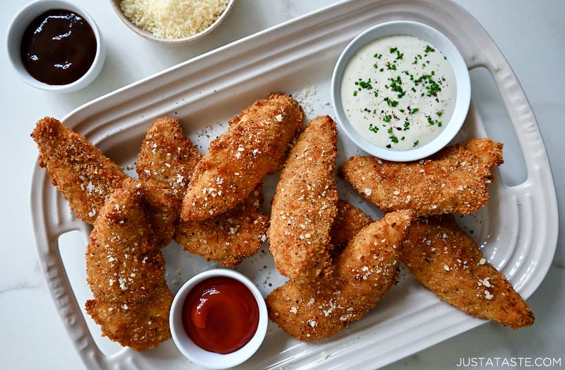 A pile of Parmesan Baked Chicken Tenders garnished with sea salt on a white serving plate with two small bowls containing Ranch dressing and ketchup