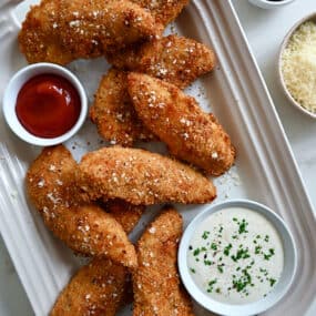 A top-down view of Parmesan Baked Chicken Tenders on a serving platter next to small bowls containing ranch dressing and ketchup