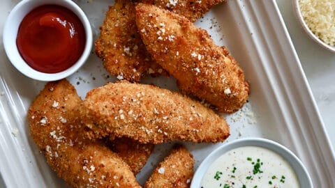 A top-down view of Parmesan Baked Chicken Tenders on a serving platter next to small bowls containing ranch dressing and ketchup