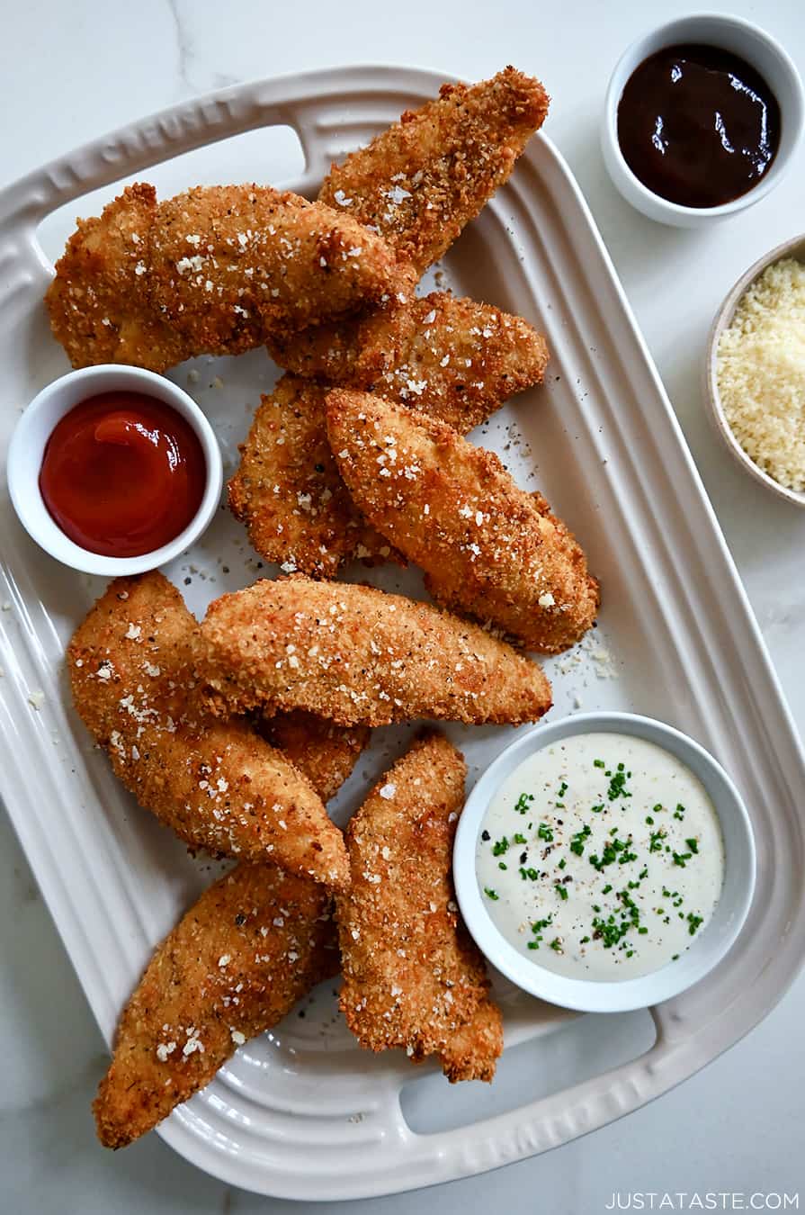 A top-down view of Parmesan Baked Chicken Tenders on a serving platter next to small bowls containing ranch dressing and ketchup