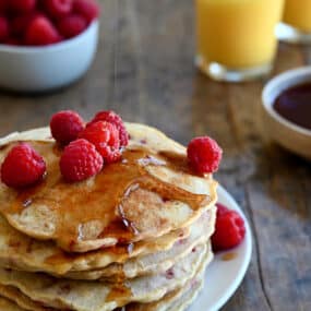 A tall stack of Raspberry Oatmeal Pancakes topped with maple syrup and fresh raspberries