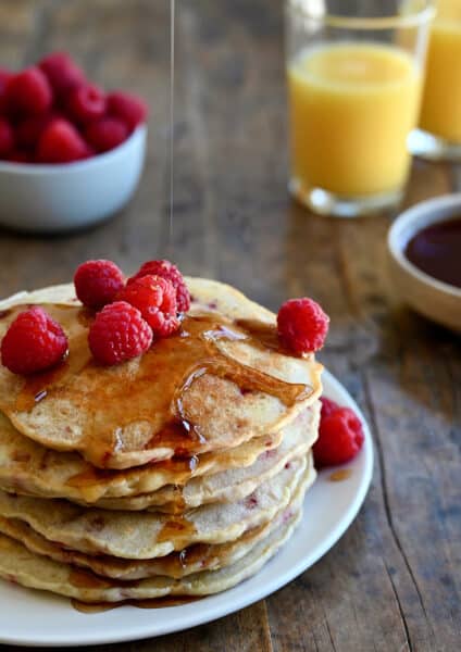 A tall stack of Raspberry Oatmeal Pancakes topped with maple syrup and fresh raspberries
