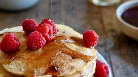 A tall stack of Raspberry Oatmeal Pancakes topped with maple syrup and fresh raspberries