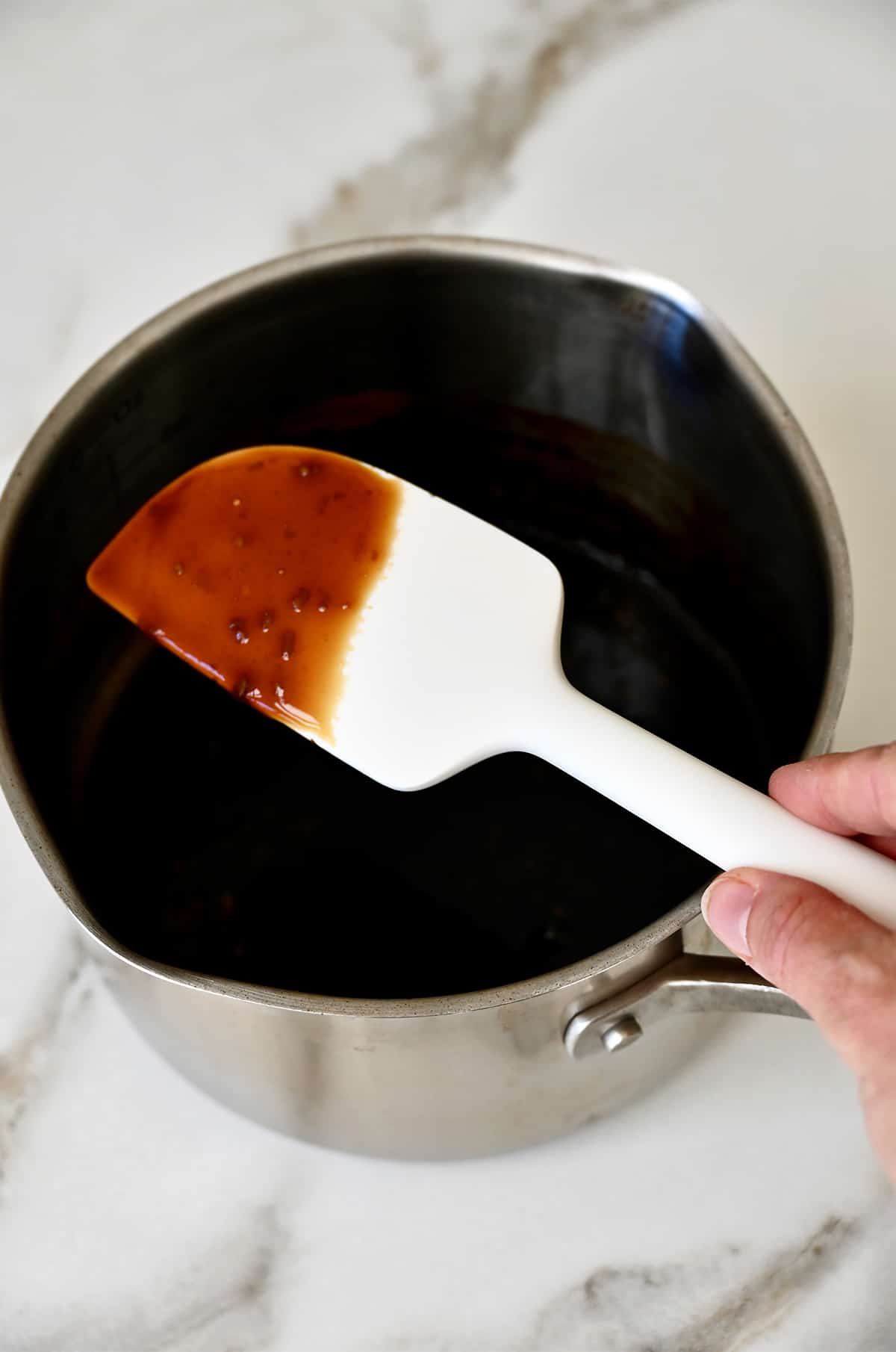 A hand holds a spatula covered in homemade teriyaki sauce over a small saucepan.