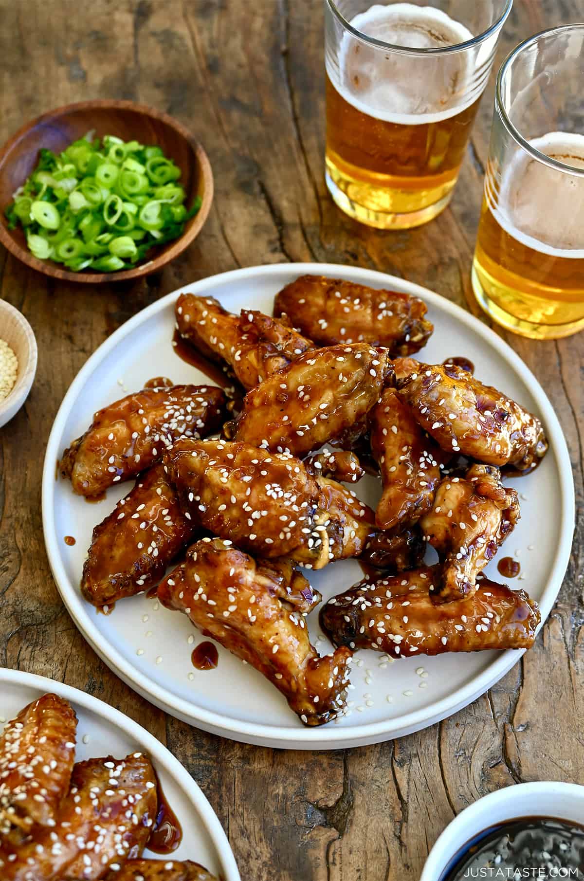 Baked chicken wings coated in teriyaki sauce and sprinkled with sesame seeds on a white plate next to two glasses containing beer and a small bowl with sliced scallions.