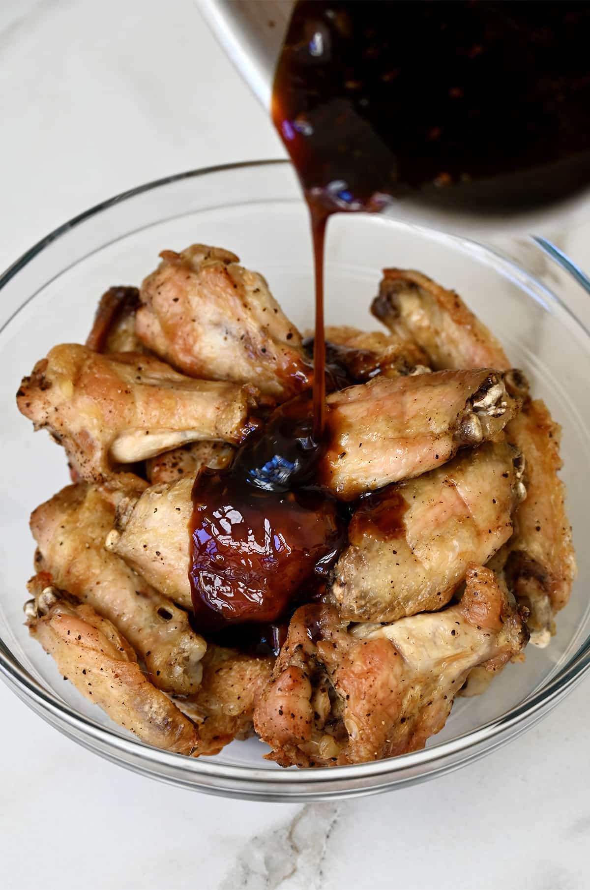 Teriyaki glaze being poured from a saucepan over baked chicken wings in a glass bowl.