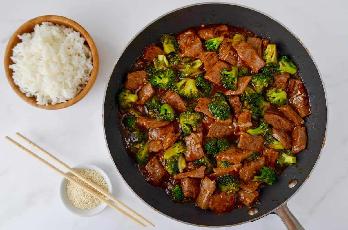 Stir-fried beef and broccoli in a garlicky soy sauce in a nonstick skillet next to a small bowl containing sesame seeds.