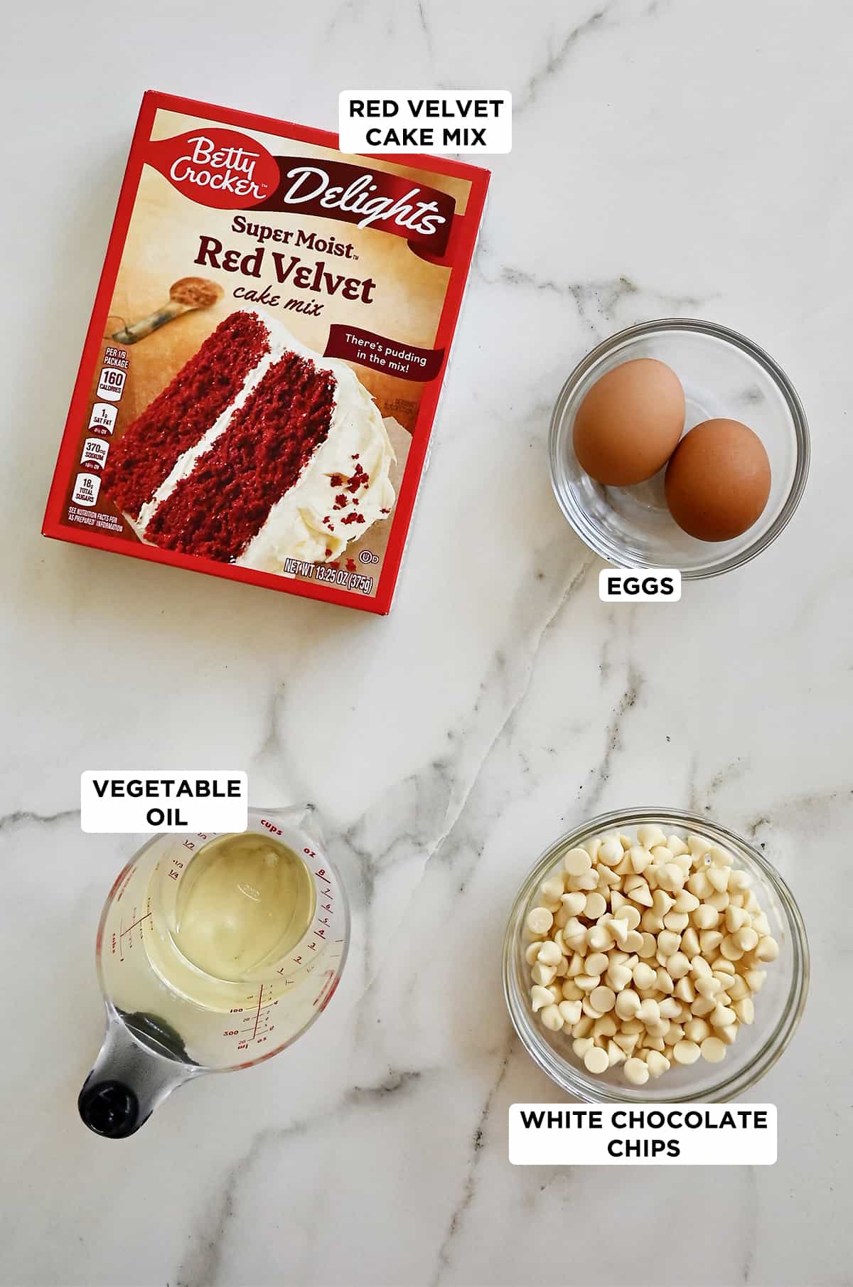 A box of red velvet cake mix is next to a small bowl containing two eggs, a small bowl containing white chocolate chips, and a liquid measuring cup filled with vegetable oil.