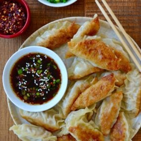 Plate containing easy chicken potstickers next to small bowl with soy dipping sauce and chopsticks next to small bowls with chopped scallions and red pepper flakes