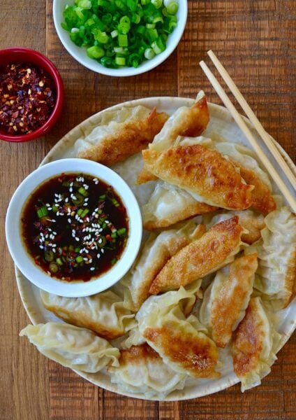 Plate containing easy chicken potstickers next to small bowl with soy dipping sauce and chopsticks next to small bowls with chopped scallions and red pepper flakes