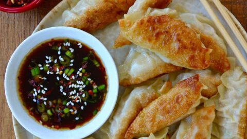 Plate containing easy chicken potstickers next to small bowl with soy dipping sauce and chopsticks next to small bowls with chopped scallions and red pepper flakes